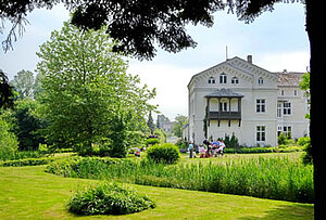 Foto eines sommerlichen Schlossparks mit Rasen und Bäumen von Schloss Roggow, einer weißen Fassade, bei schönem Wetter